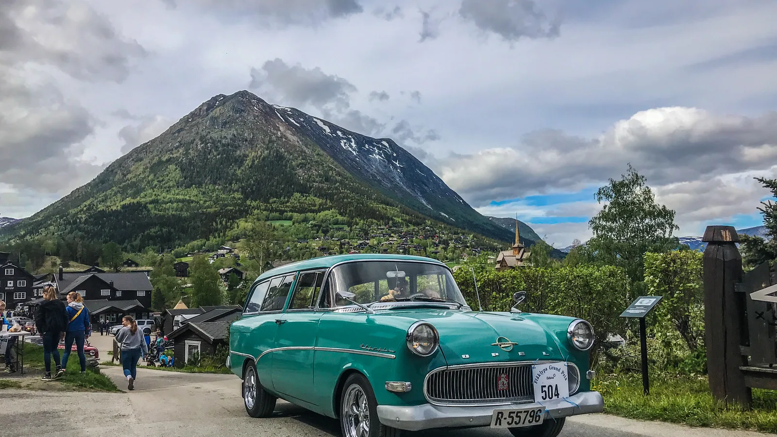 A vintage turquoise Opel station wagon at the Flåklypa Grand Prix rally in Lom, with scenic mountain backdrop and cheering spectators