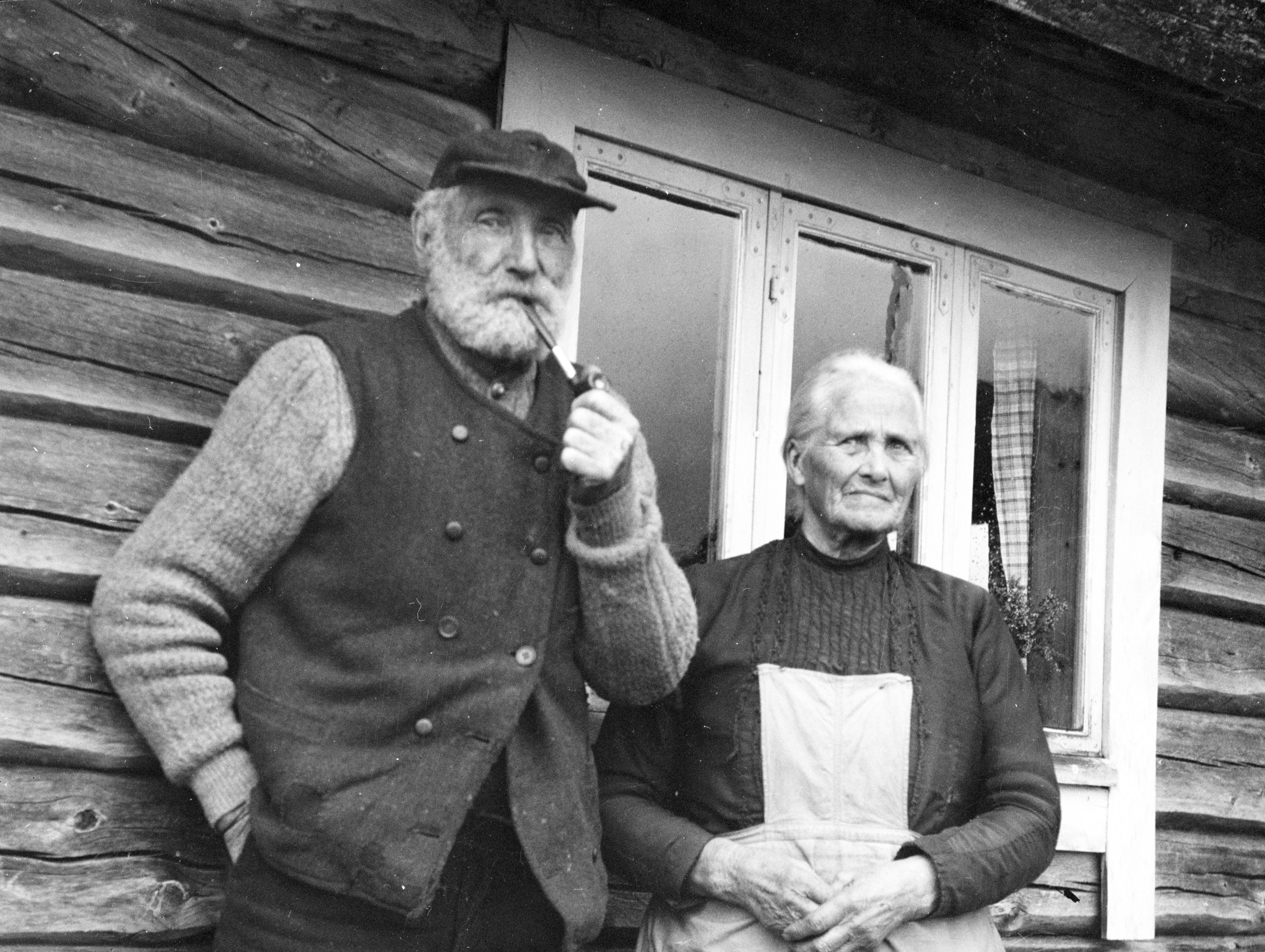 A black-and-white portrait of Reodor and Kristine Wardenær in front of a log cabin at Fiskevollen — Reodor with pipe and flat cap, Kristine in apron