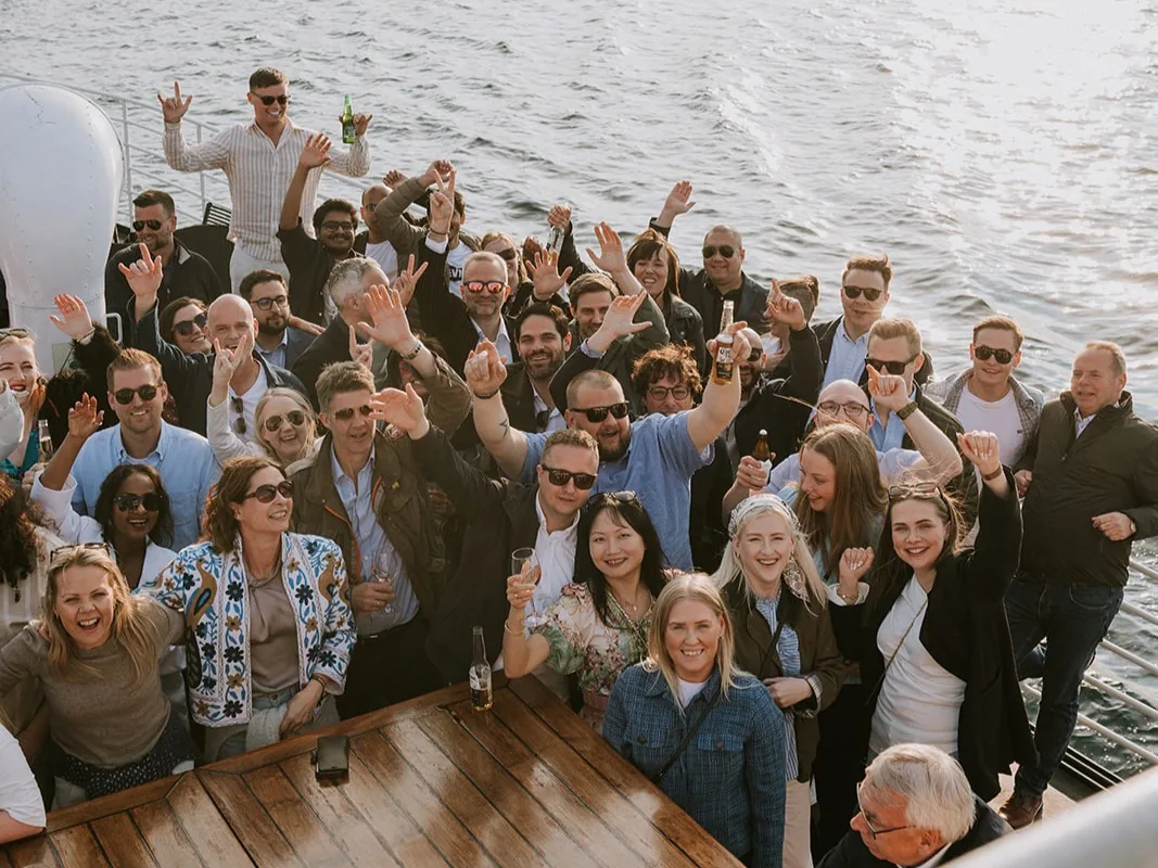 A large group of smiling VARD employees standing on the deck of a boat at sea, raising their hands and enjoying a social event together.