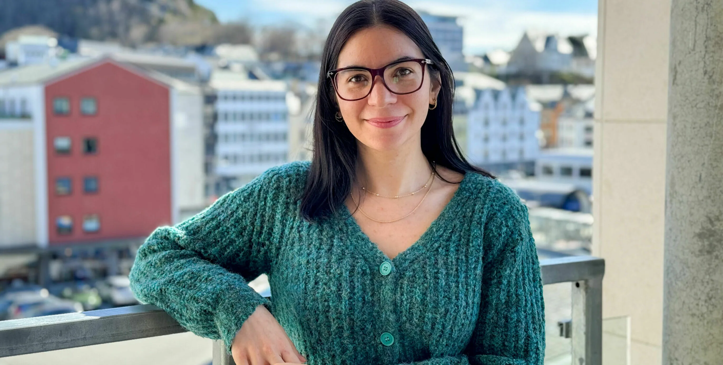 Gema Baldo Llorca leaning on a balcony railing with the colourful buildings of Ålesund and mountains in the background.