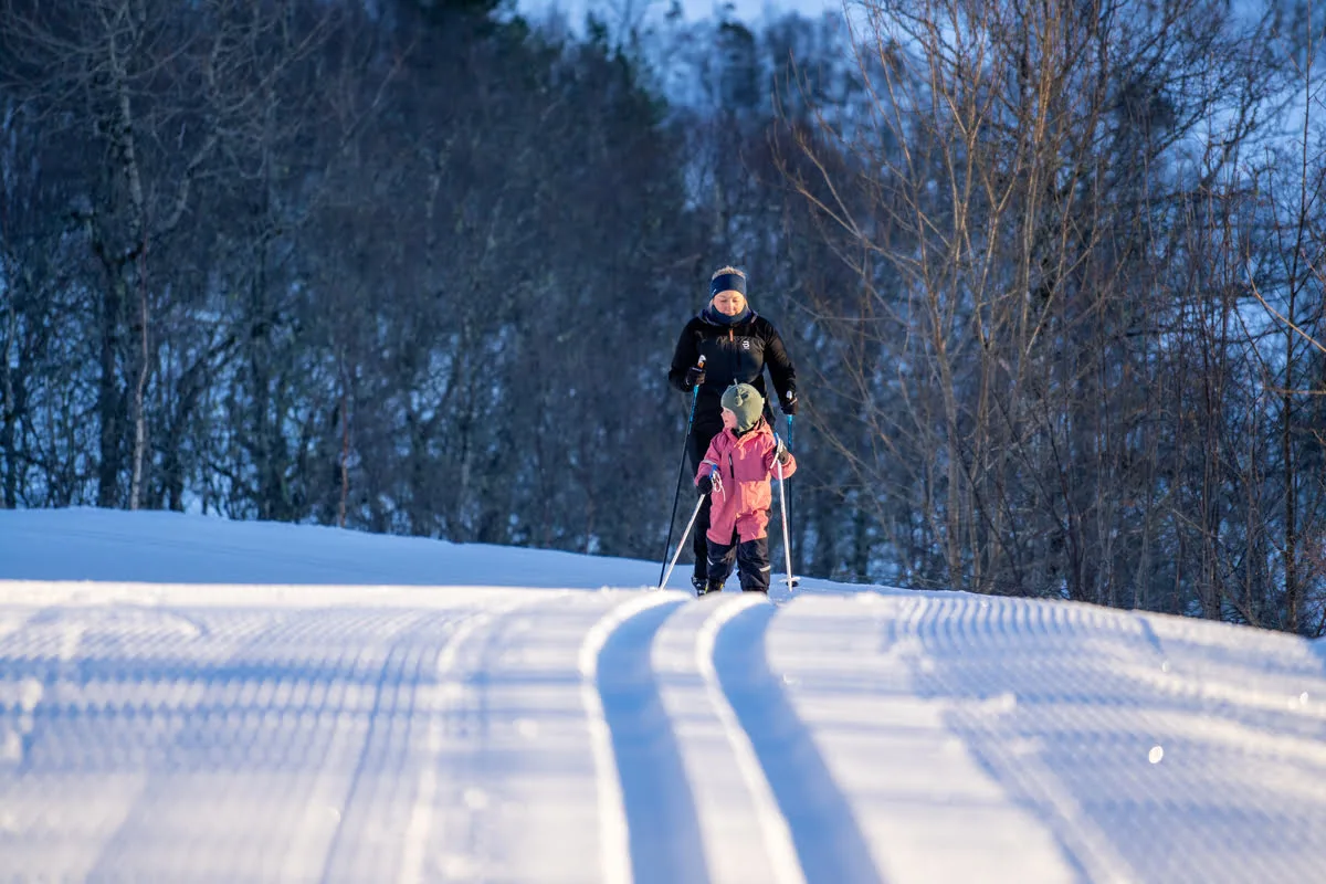 Adult and child cross-country skiing on freshly groomed tracks in Tomrefjord, with snow and forest in the background